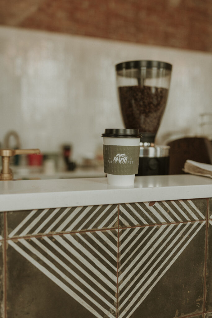 A branded Faire Coffee to-go cup resting on a modern coffee bar in Eureka, IL, surrounded by coffee syrups and equipment, showcasing a clean and inviting café aesthetic.