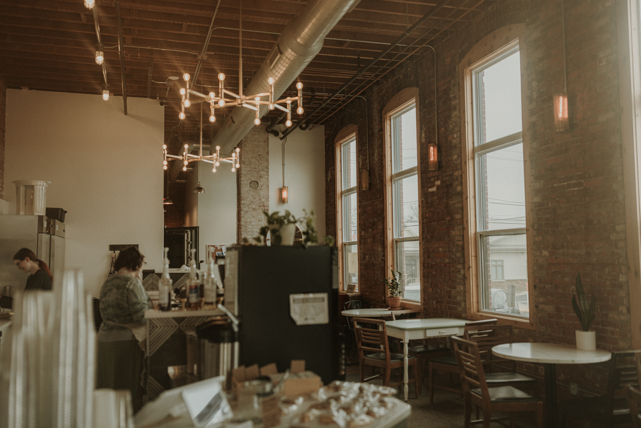 A warmly lit coffee shop in Eureka, IL, featuring exposed brick walls, large windows letting in natural light, and cozy tables with potted plants. The barista team is seen working behind the counter, creating a welcoming atmosphere.